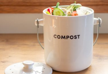 A white compost container filled with vegetable scraps sits on a wooden kitchen counter beside its lid.