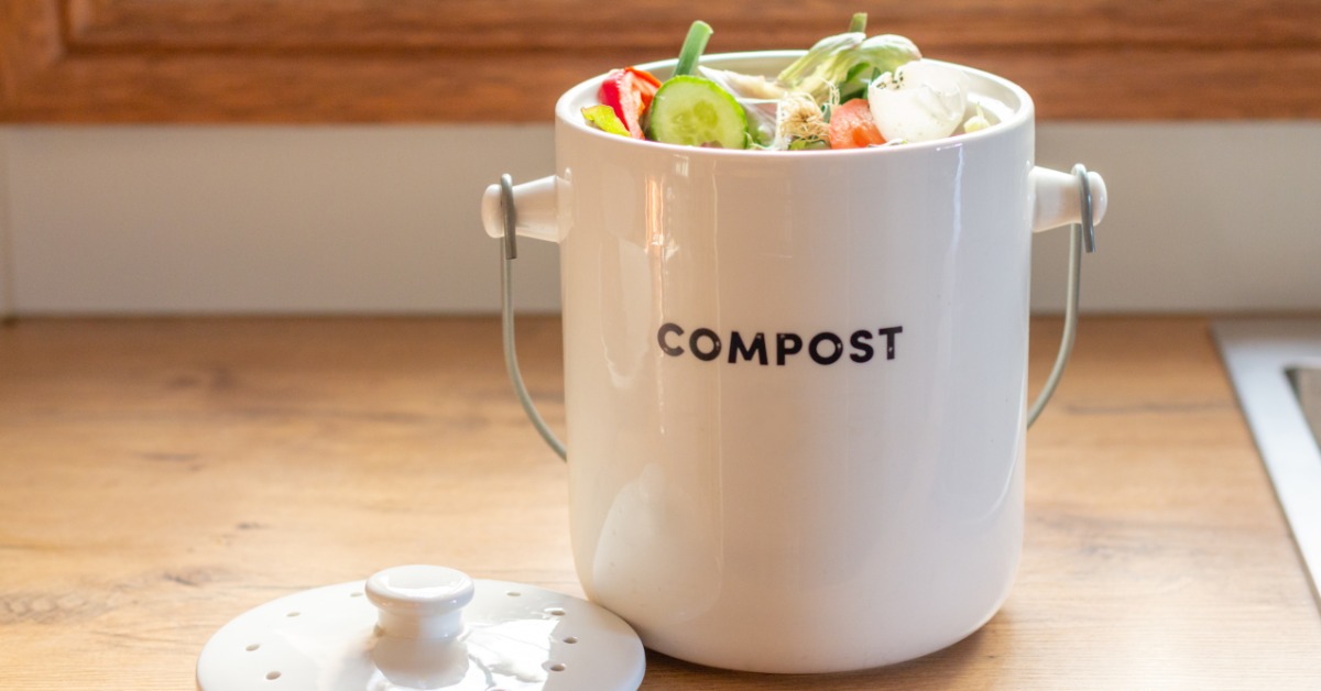 A white compost container filled with vegetable scraps sits on a wooden kitchen counter beside its lid.
