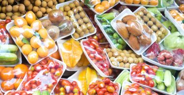 Trays of wrapped fruits and vegetables, including tomatoes, cucumbers, mangoes, and longans, are on display