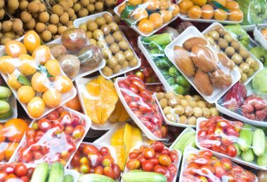 Trays of wrapped fruits and vegetables, including tomatoes, cucumbers, mangoes, and longans, are on display