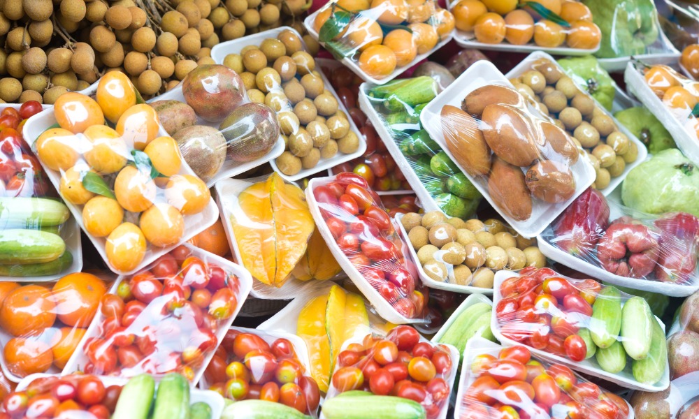 Trays of wrapped fruits and vegetables, including tomatoes, cucumbers, mangoes, and longans, are on display
