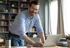 An entrepreneur stands over his desk with his hand on his laptop and he smiles. His desk is in an office.