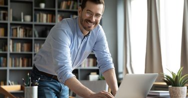 An entrepreneur stands over his desk with his hand on his laptop and he smiles. His desk is in an office.