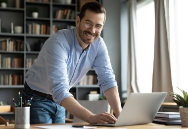 An entrepreneur stands over his desk with his hand on his laptop and he smiles. His desk is in an office.