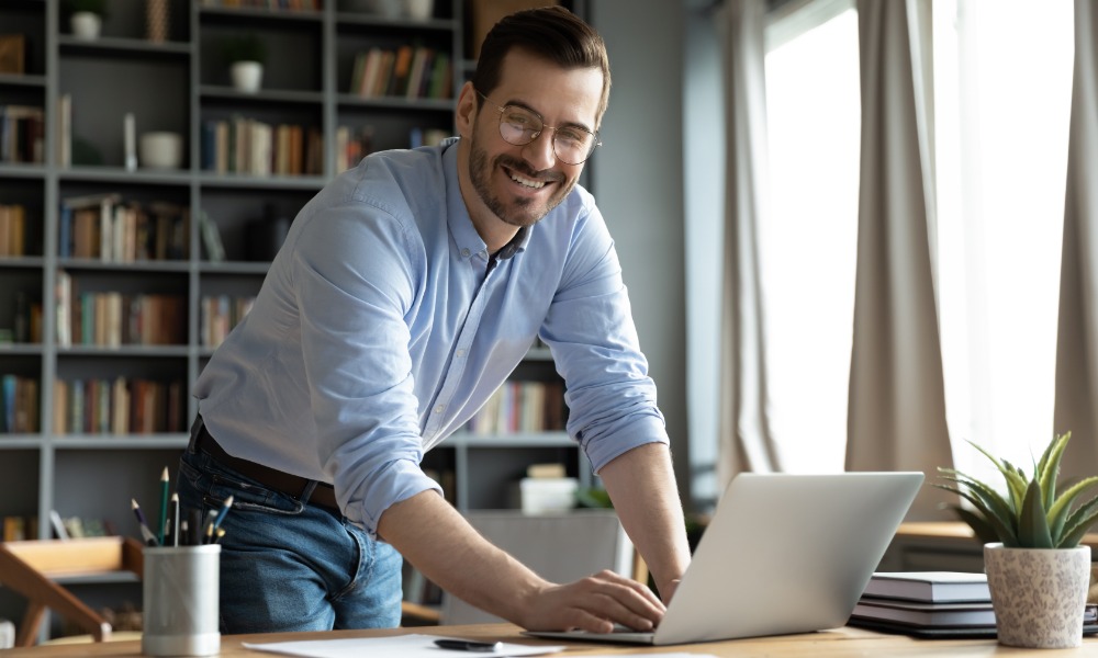 An entrepreneur stands over his desk with his hand on his laptop and he smiles. His desk is in an office.