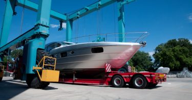 White motorboat rests on a red transport trailer beneath large blue boat-lifting cranes at a marina.