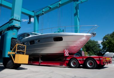 White motorboat rests on a red transport trailer beneath large blue boat-lifting cranes at a marina.