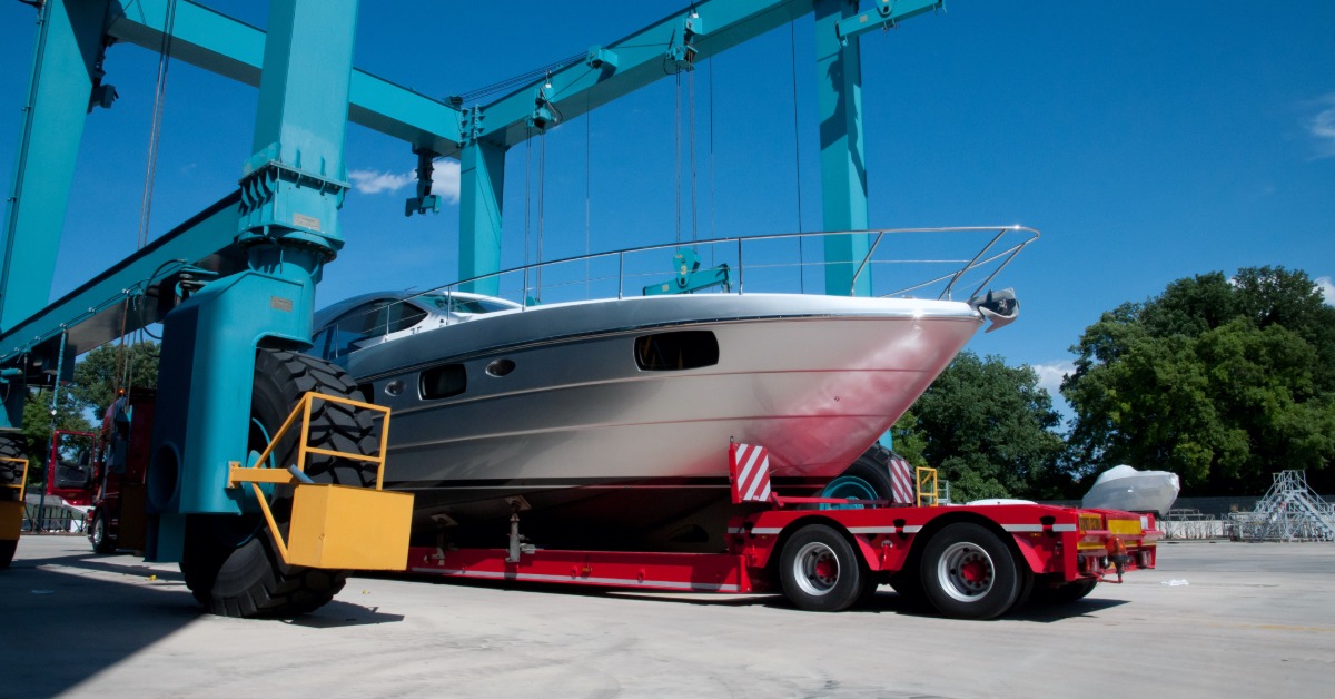 White motorboat rests on a red transport trailer beneath large blue boat-lifting cranes at a marina.