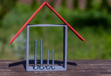 A small house-shaped model with a red roof and four silver screws stands on a wooden surface outdoors.