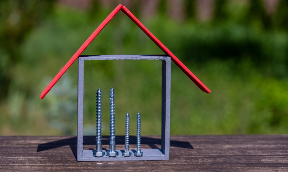 A small house-shaped model with a red roof and four silver screws stands on a wooden surface outdoors.