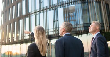 Three people in suits standing near a glass building, with one person pointing toward upper windows.