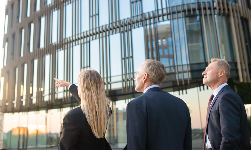 Three people in suits standing near a glass building, with one person pointing toward upper windows.