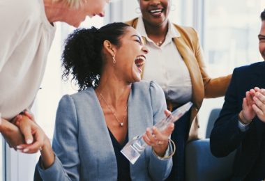 An employee who just received an award is happy and laughing as her coworkers cheer along with her next to her.