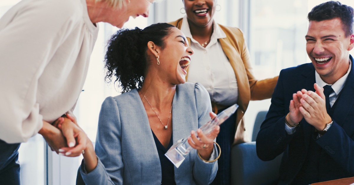 An employee who just received an award is happy and laughing as her coworkers cheer along with her next to her.