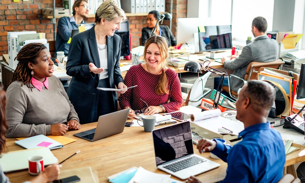A group of people gathered around desks with laptops, papers, coffee mugs, and office shelves behind them.