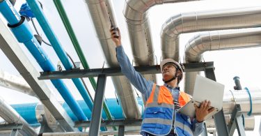 A low-angle view of a worker in an orange safety vest holding a laptop and radio below metal pipes outdoors.