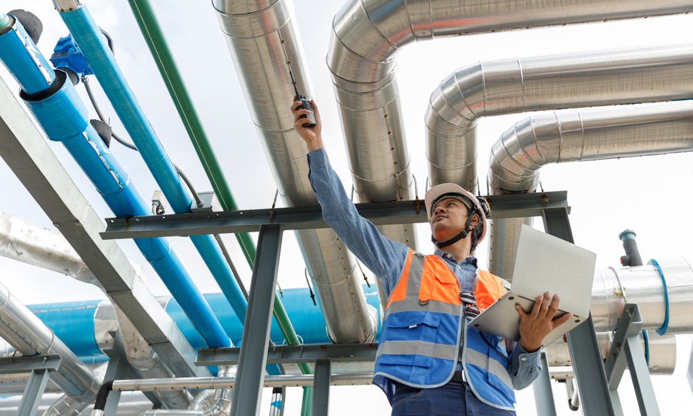 A low-angle view of a worker in an orange safety vest holding a laptop and radio below metal pipes outdoors.