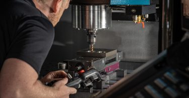 A person holding a tool beside a CNC machine spindle that's drilling into a metal block inside dark equipment.