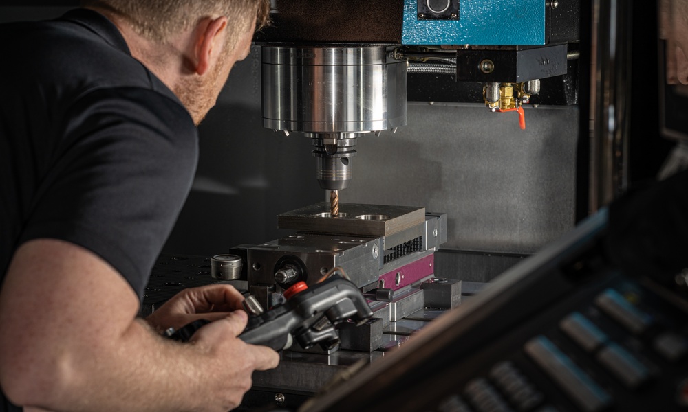 A person holding a tool beside a CNC machine spindle that's drilling into a metal block inside dark equipment.