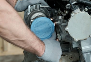 A person wearing gloves positions a silver turbocharger into a car engine, with hoses and metal parts nearby.