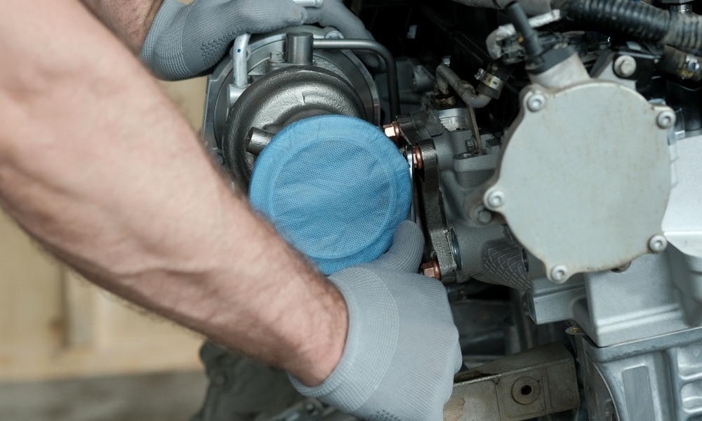 A person wearing gloves positions a silver turbocharger into a car engine, with hoses and metal parts nearby.