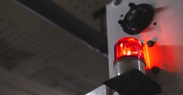 A red warning light glows on a metal surface in a dim industrial area, surrounded by bolts and shadows.