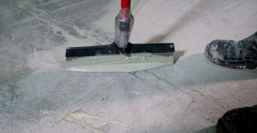 A close-up of a squeegee tool applying epoxy resin coating to a concrete floor next to a pair of dirty boots.