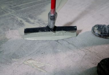 A close-up of a squeegee tool applying epoxy resin coating to a concrete floor next to a pair of dirty boots.