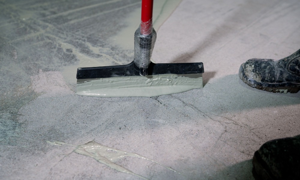 A close-up of a squeegee tool applying epoxy resin coating to a concrete floor next to a pair of dirty boots.