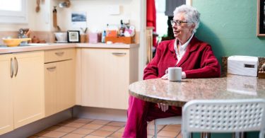A senior woman wearing a pink corduroy suit, sitting inside a kitchen with green walls next to a white cup on a table.