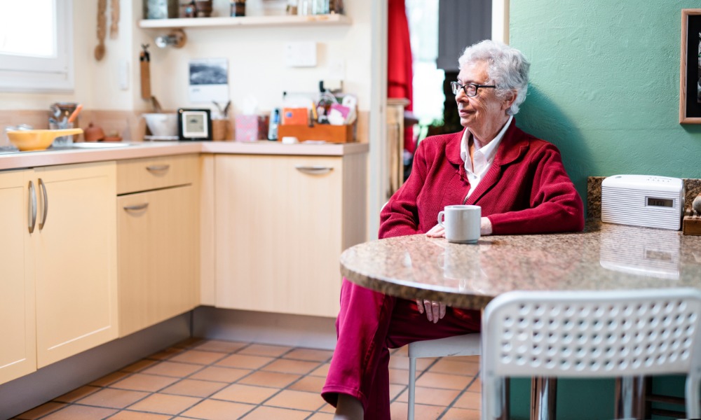 A senior woman wearing a pink corduroy suit, sitting inside a kitchen with green walls next to a white cup on a table.