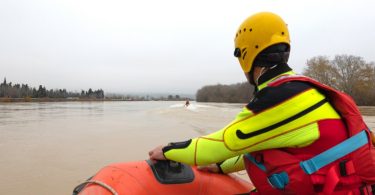 A person wearing a brightly colored wetsuit, a yellow helmet, and a red life jacket is riding in a red rescue boat.