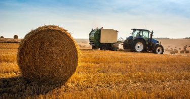 A close-up of a round hay bale in a hay field with a blue tractor towing a round hay baler in the background.