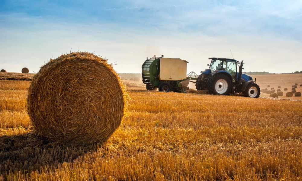 A close-up of a round hay bale in a hay field with a blue tractor towing a round hay baler in the background.