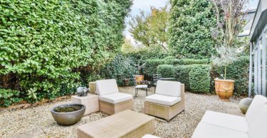 A backyard covered in gravel and surrounded by tall bushes and trees, with outdoor couches and a teak coffee table.