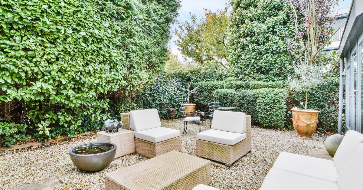A backyard covered in gravel and surrounded by tall bushes and trees, with outdoor couches and a teak coffee table.