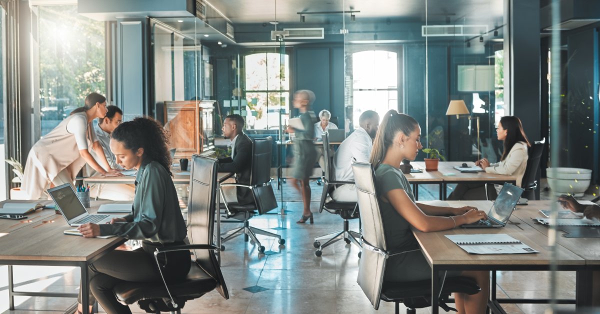 A large office space shows many people sitting at desks and working at computers. Other people are walking around.