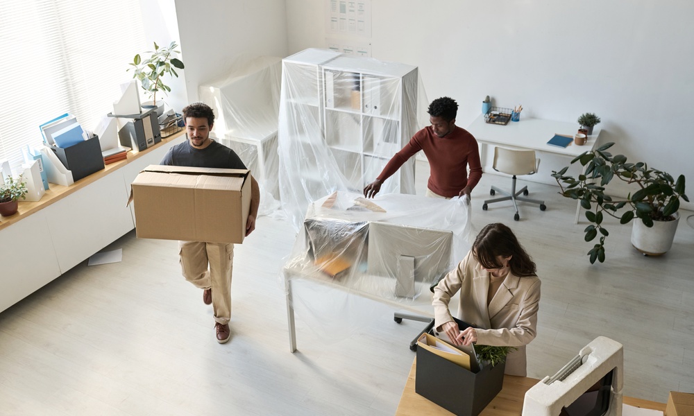 Three people are packing things up in an office environment. There is a window to the left of frame.