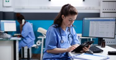 A female nurse is sitting at a desk and is holding a tablet in her hands. There is another woman seated behind her.