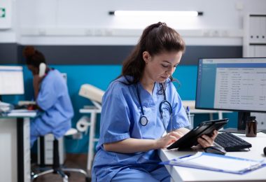 A female nurse is sitting at a desk and is holding a tablet in her hands. There is another woman seated behind her.