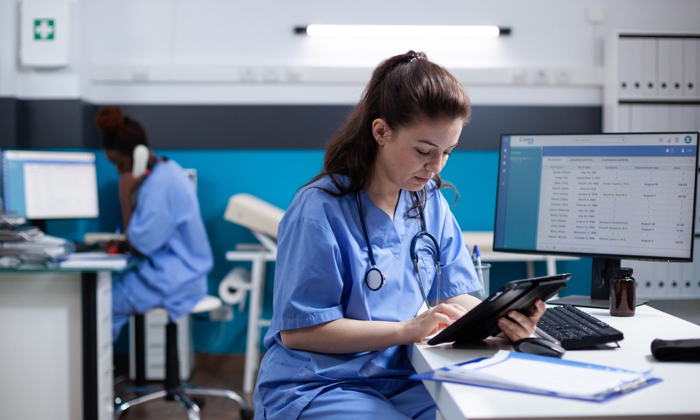 A female nurse is sitting at a desk and is holding a tablet in her hands. There is another woman seated behind her.