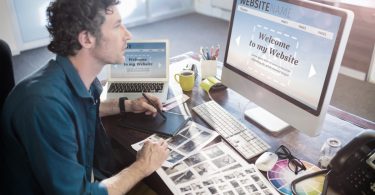 Designer works at a desk with a graphics tablet, color swatches, printed layouts, and a website open on a desktop.