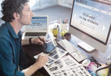 Designer works at a desk with a graphics tablet, color swatches, printed layouts, and a website open on a desktop.