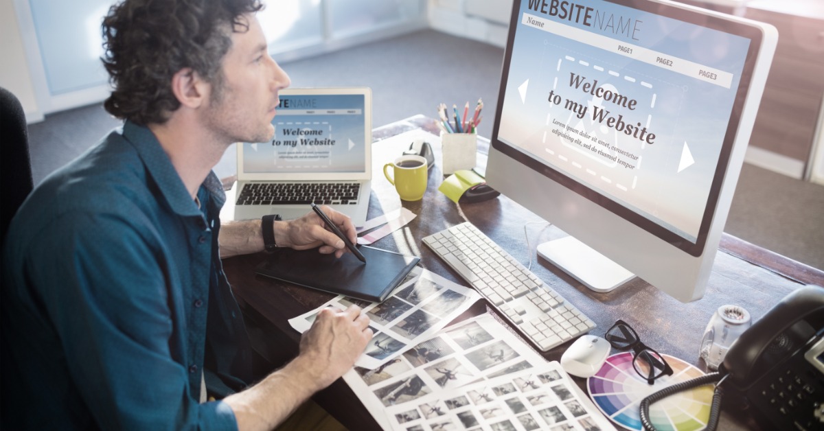 Designer works at a desk with a graphics tablet, color swatches, printed layouts, and a website open on a desktop.