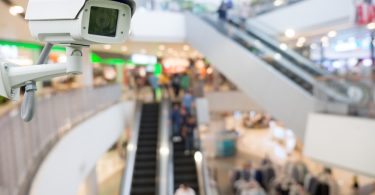 A close-up of a CCTV camera placed on the side of a wall next to multiple escalators inside a large shopping mall.