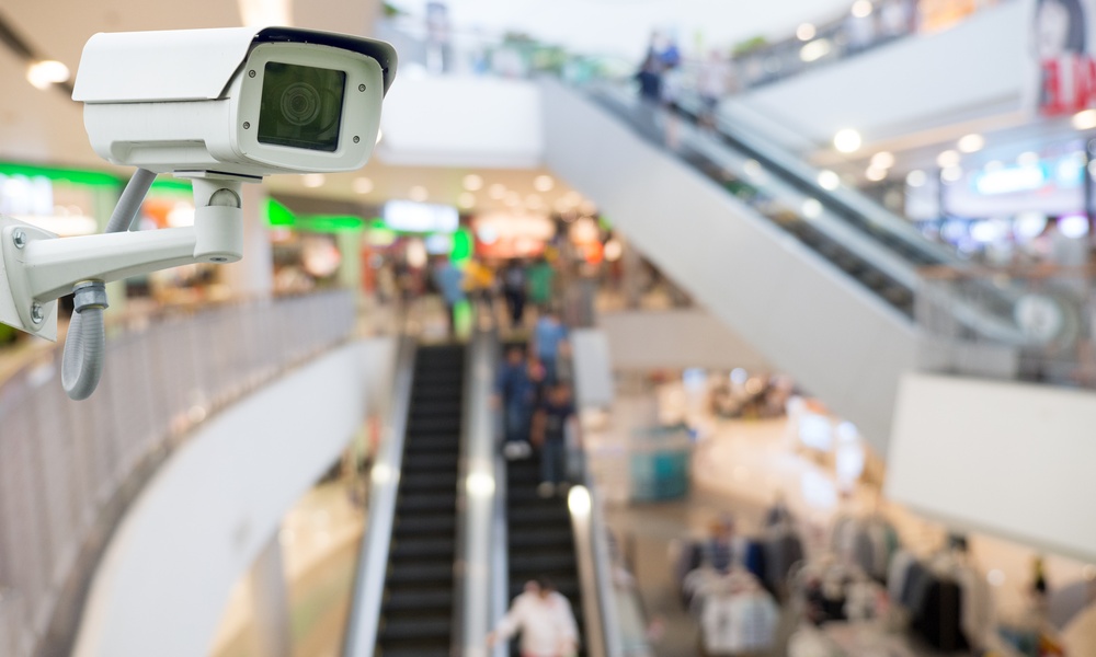 A close-up of a CCTV camera placed on the side of a wall next to multiple escalators inside a large shopping mall.