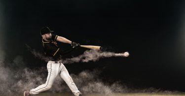 A side view of a baseball player in a black jersey with white pants swinging and contacting a baseball.
