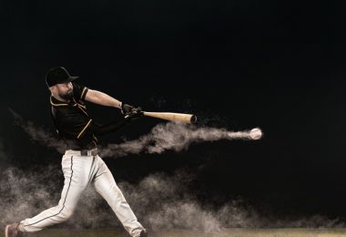 A side view of a baseball player in a black jersey with white pants swinging and contacting a baseball.