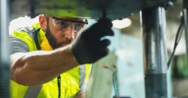 A man in a hard hat and protective eyewear is bending over and looking into a piece of industrial machinery.