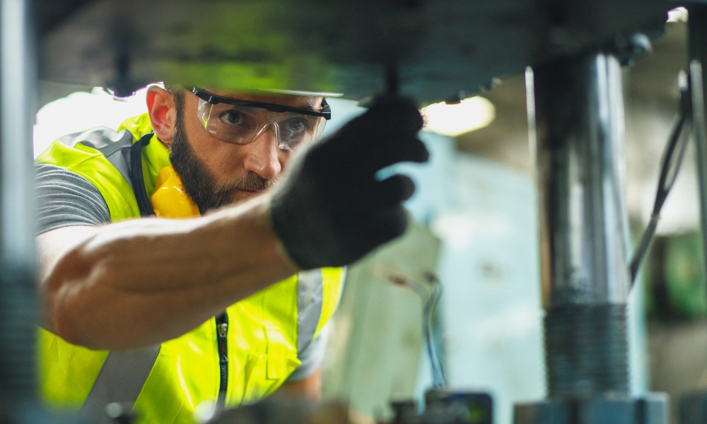 A man in a hard hat and protective eyewear is bending over and looking into a piece of industrial machinery.